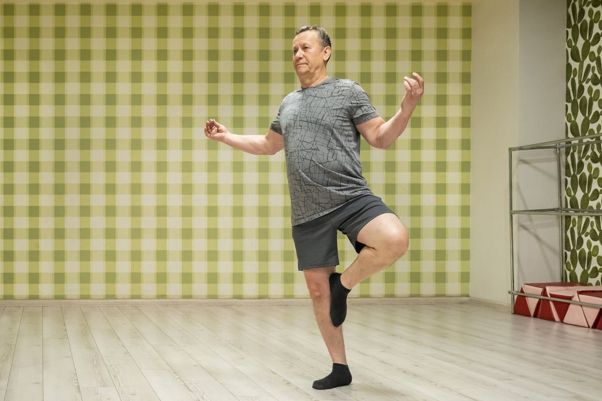 Holding tree pose adult caucasian man balancing in indoor fitness studio facing mirror improving stability concentration and body alignment after work